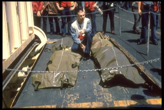 Crew member kneeling beside four body bags on ship elevator